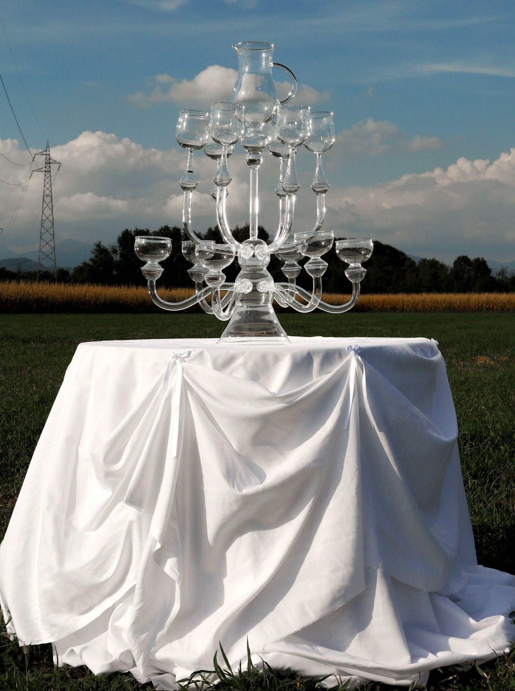 A "Dinner's Ready" Glass Sculpture by Ylaria Pavone is centered on a table draped with a white cloth. The table is set outdoors in a green field with distant trees and a cloudy sky. An electricity pylon is visible in the background. The sculpture holds several tiers of empty glasses, offering a unique dining experience with elegant glass dinnerware.