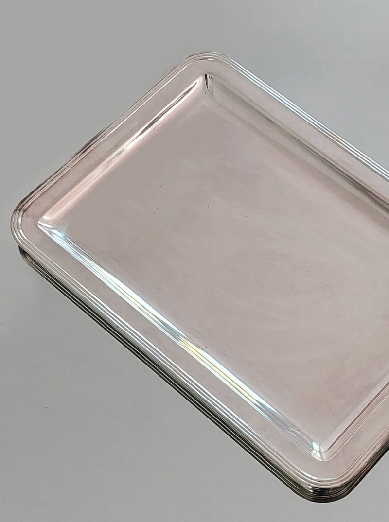 A Silver-plated Rectangle Tray from Les Objoies is placed against a plain grey background. This minimalist tray features polished edges and a smooth, reflective surface, boasting a modern aesthetic with slightly raised, rounded edges.