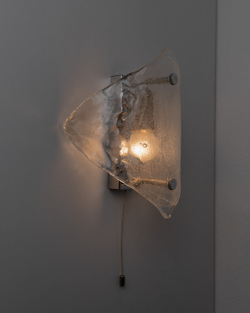 The Vintage Murano Glass Wall Lamp by Kalmar from Bottega Jacobs features a space-age design with an abstract, translucent glass shade. A warm glow emanates from the bulb held by two metal rods, and it's operated via a pull cord, embodying the elegance of handmade glass art.