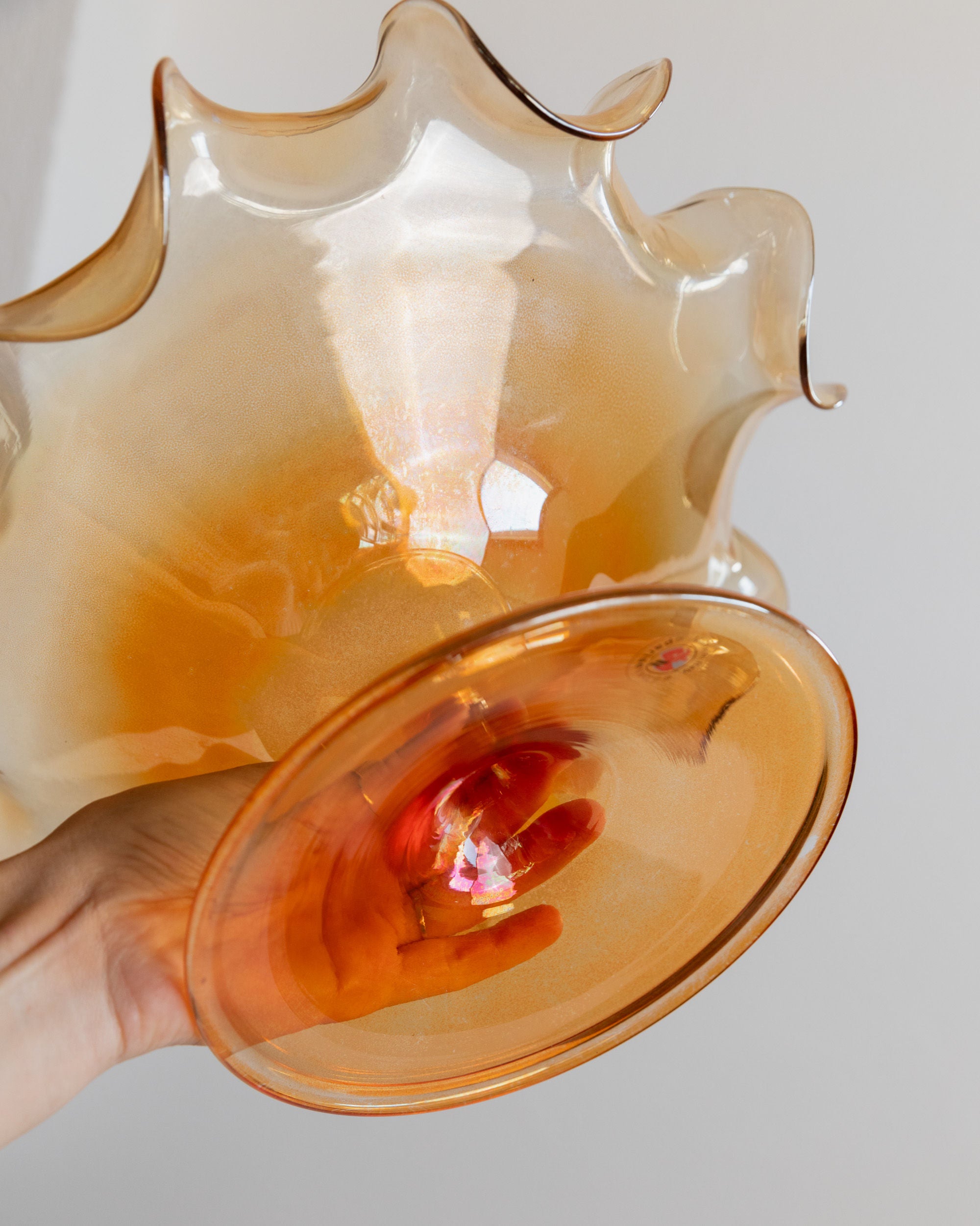 A hand holds the Murano Glass Bowl by Parise Vetro 70s from Bottega Jacobs, showcasing its amber-colored, wavy design with a smooth, wide base. The light shines through, creating a warm glow reminiscent of orange iridescence. The softly blurred background accentuates the intricate design and texture of this captivating decorative piece.