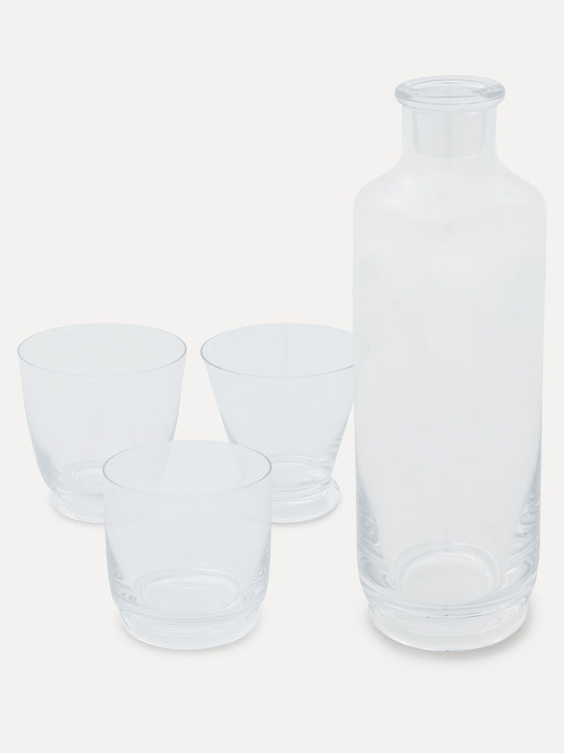 A clear glass carafe stands next to four Walter 01 Tumblers from Service Projects, all crafted from lead-free crystal. The set is placed against a plain, light-colored background. The carafe has a simple, cylindrical shape with a slightly narrower neck, and the tumblers are round with smooth edges, exuding subtle Art Deco elegance.