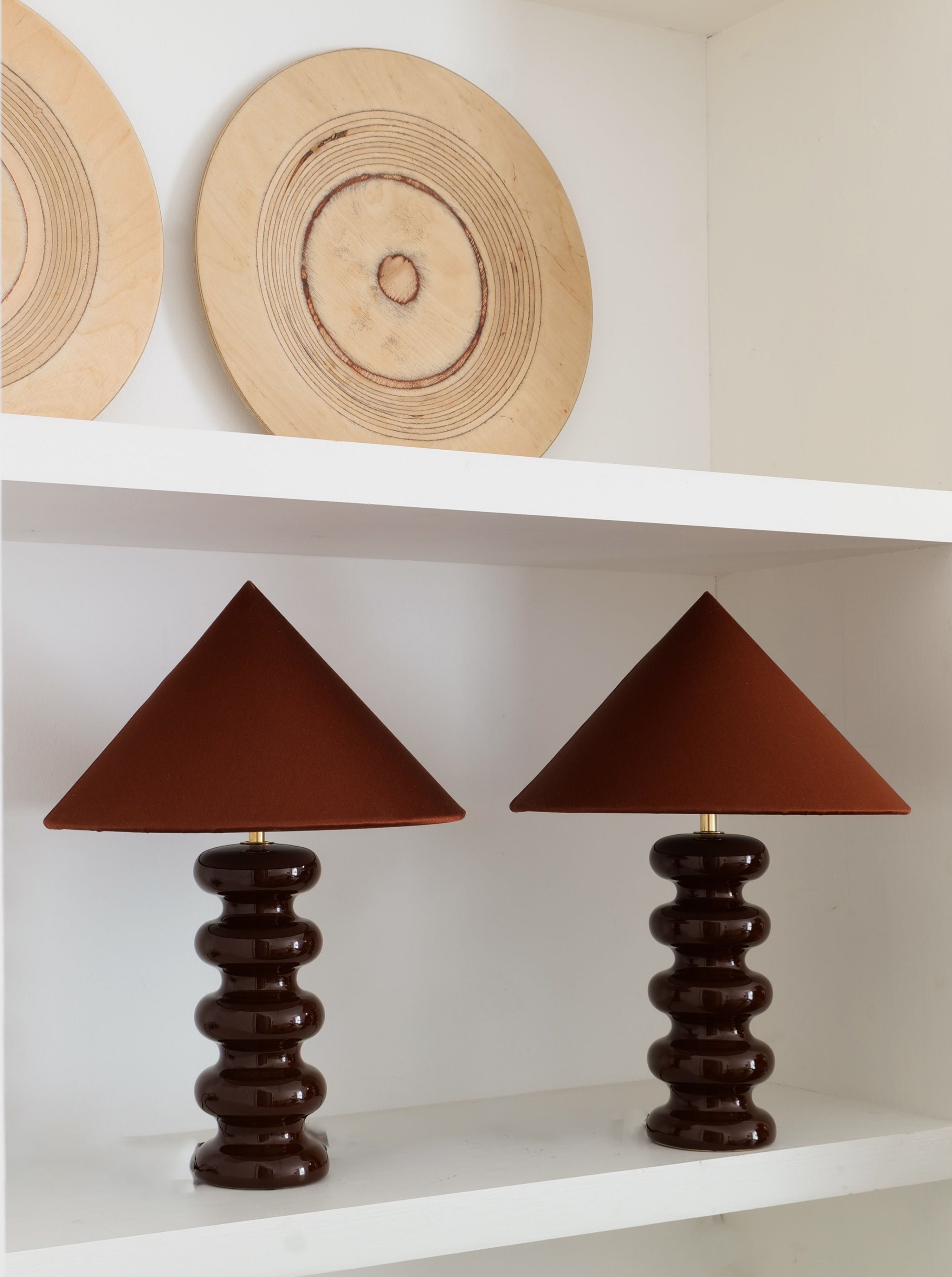 A white shelf displays a Collection apart Sculptural Ceramic Lamp beside another dark brown table lamp with a conical shade on the lower shelf. Above them, two decorative, round wooden plates with concentric circular patterns rest against the wall, creating a minimalistic and earthy aesthetic.