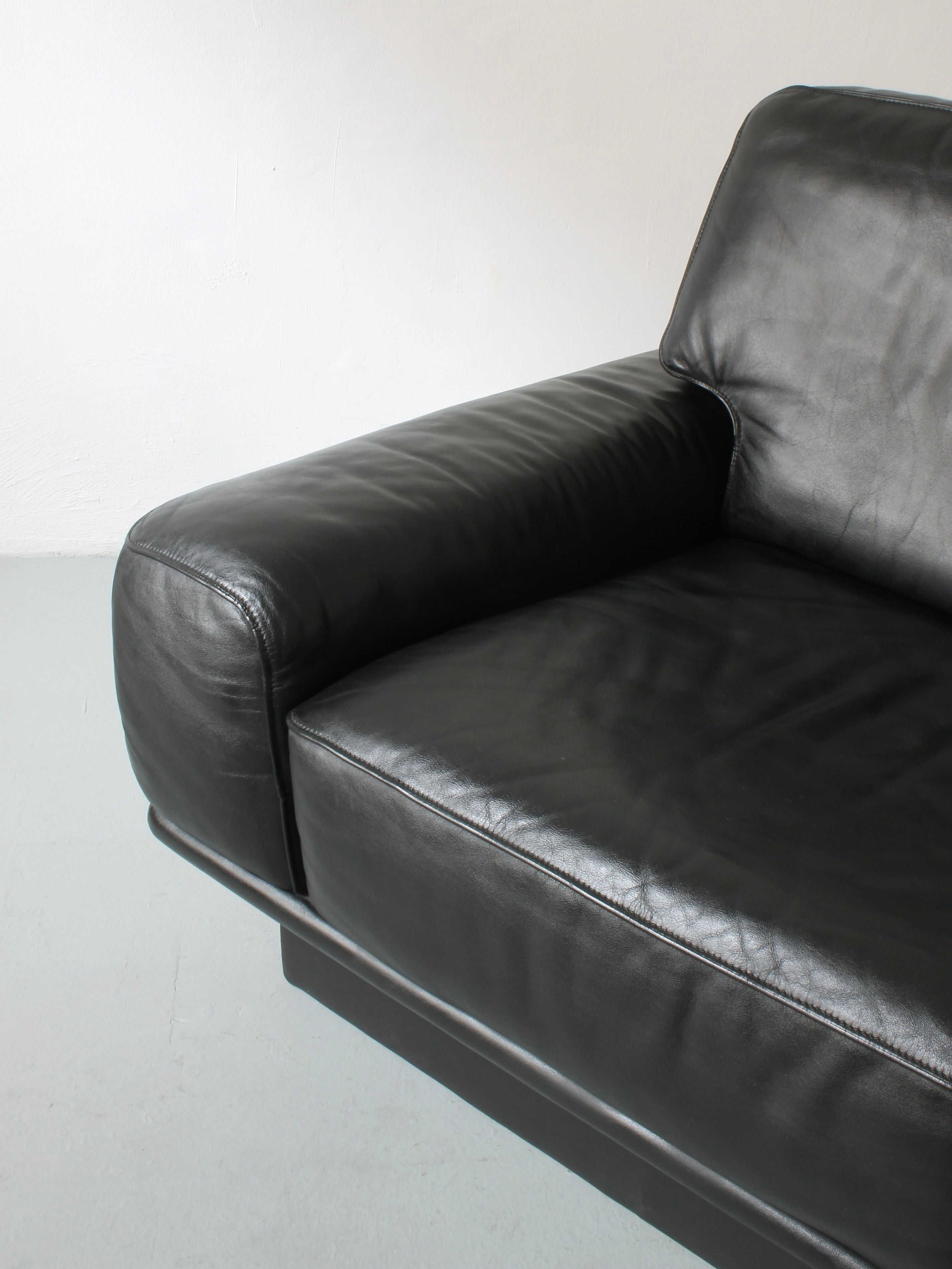 The Lounge Chair by De Sede from Afterhour Furniture is showcased, featuring Swiss luxury with its modern black leather design. The image highlights the right armrest and part of the seat, revealing the smooth texture of high-quality leather on a light-colored floor.