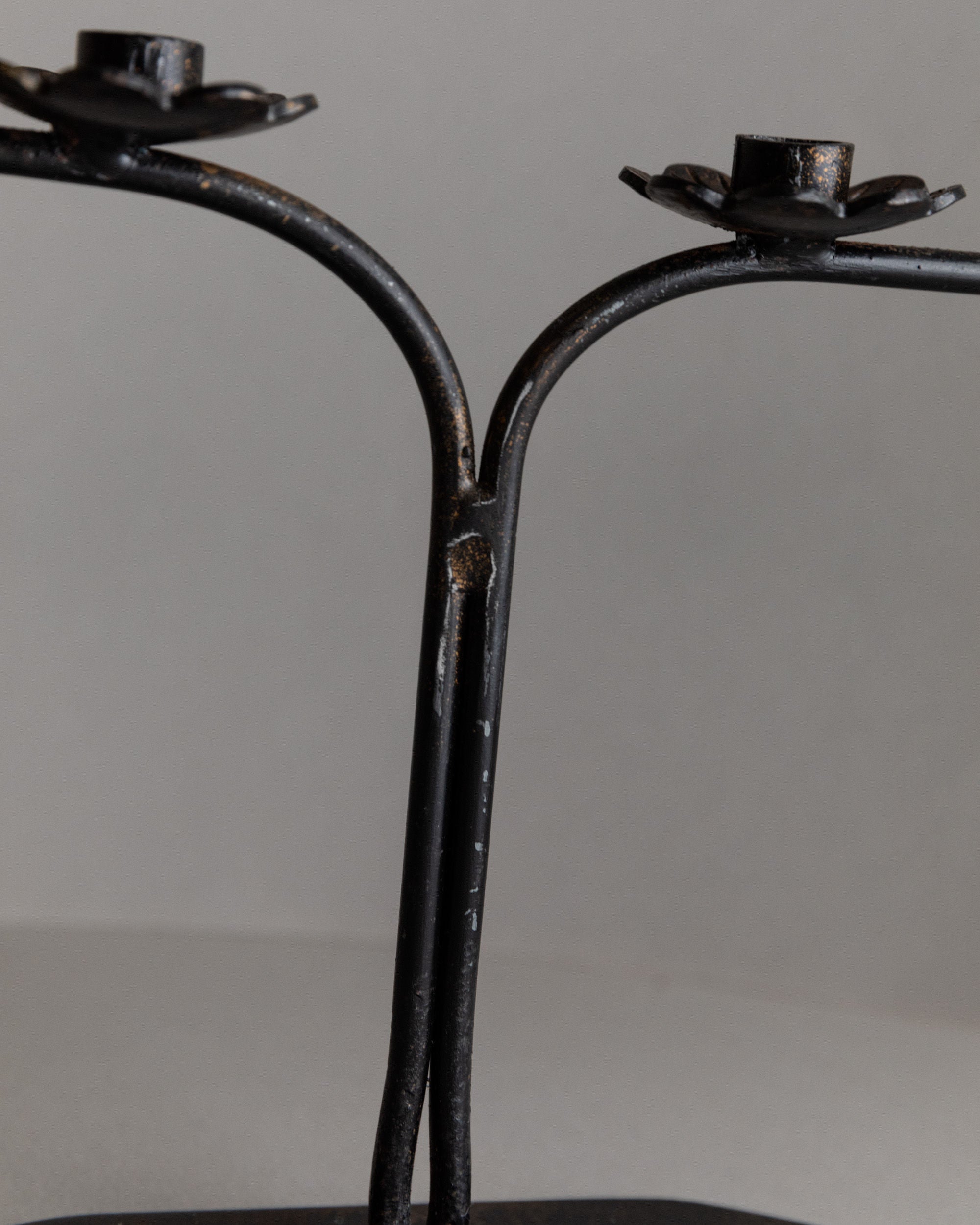 A close-up of the Iron Candleholder by Bottega Jacobs, crafted in Italy in the 1960s, showcases two branched arms ending in flower-shaped platforms for candles. Its aged, rustic finish makes it an ideal table decoration against a plain background.