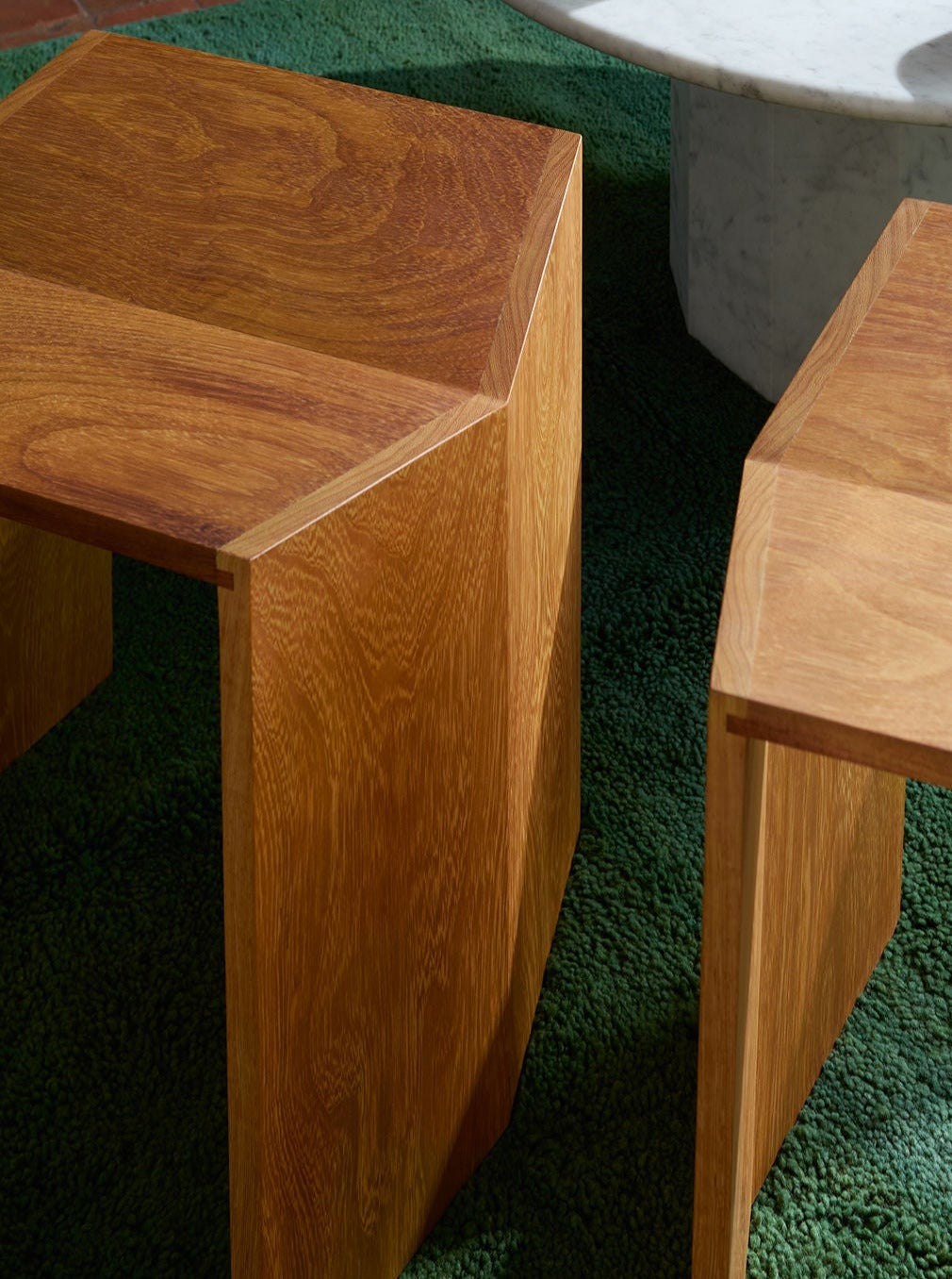 Close-up of wooden table corners on a textured green carpet, featuring smooth lines and rich wood grain detail from solid Lemon Athens Stool in Iroko, with a glimpse of a marble object in the background.