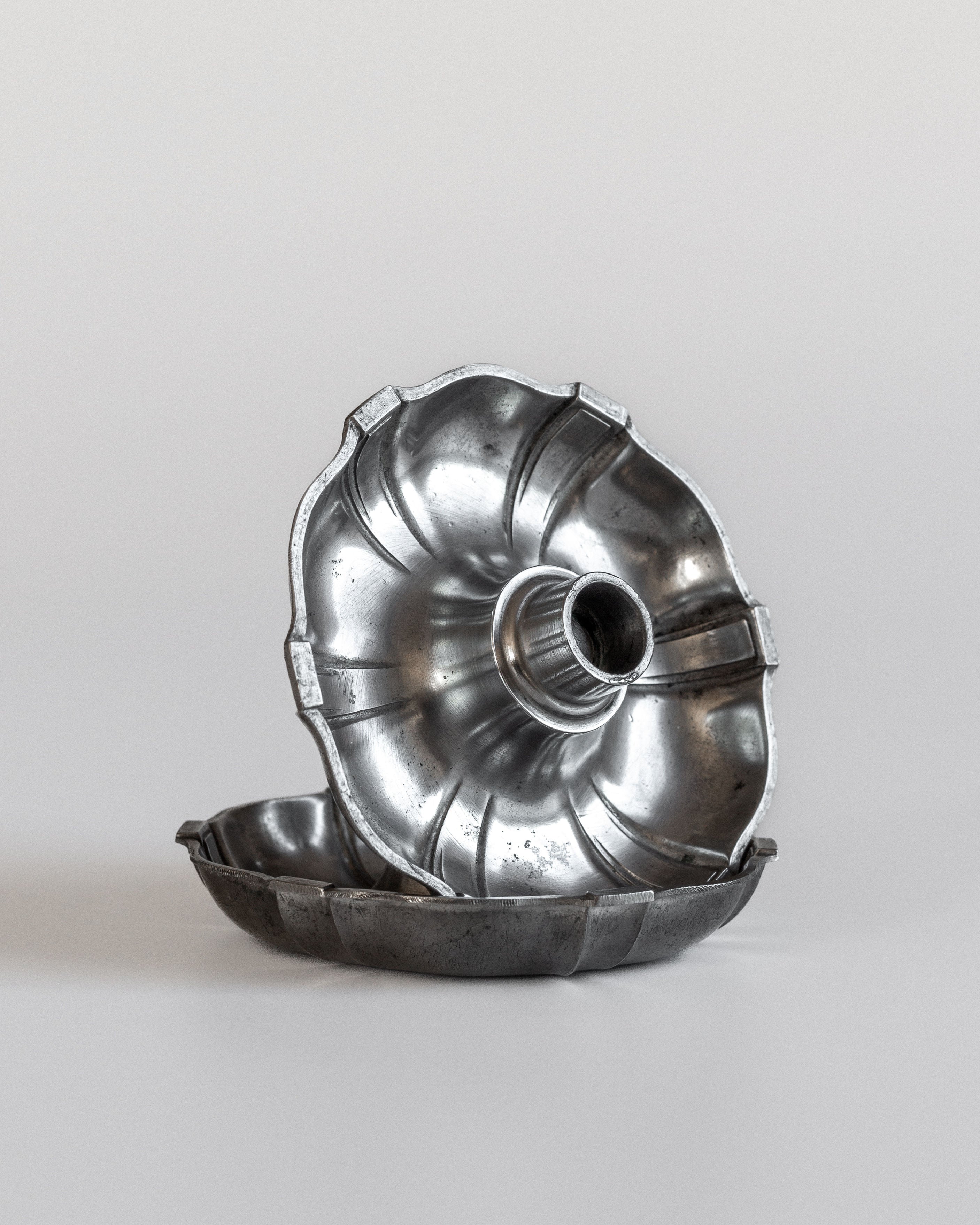 Two silver bundt cake pans displayed against a neutral background evoke the elegance of Spigel's Modernist Metal Candleholders from the 1930s. One pan is upright, showcasing its ridged interior like finely crafted candleholders, while the other lies flat, revealing its worn, metallic surface reminiscent of C.G. Hallberg's designs.