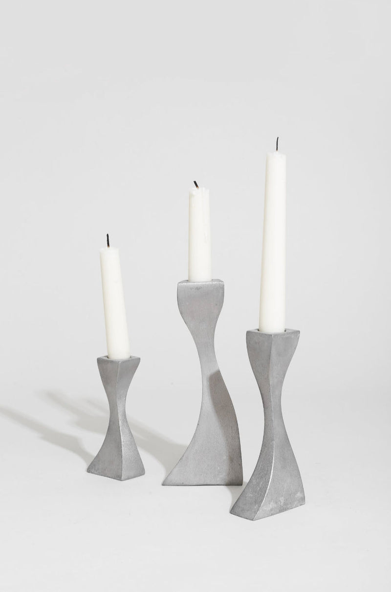 The Villa Wavy Candleholder Set features a sleek metal trio with hourglass shapes and tapered edges, displaying white candles with blackened wicks against a simple white background.