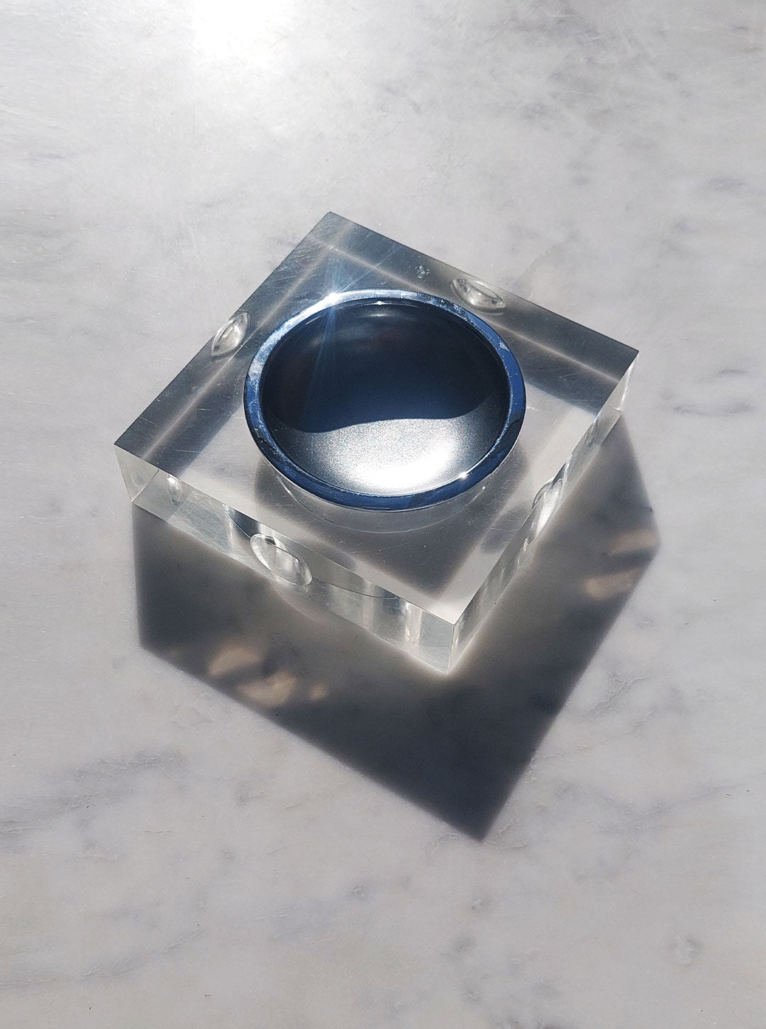 A round sculptural ring with a glossy blue surface is embedded in a clear, square resin block, reminiscent of Les Objoies' 70's Plexiglass Ashtray designs. The block sits on a light grey, marbled countertop. Sunlight creates reflections and shadows, adding a sense of depth and dimension to the minimalist design.