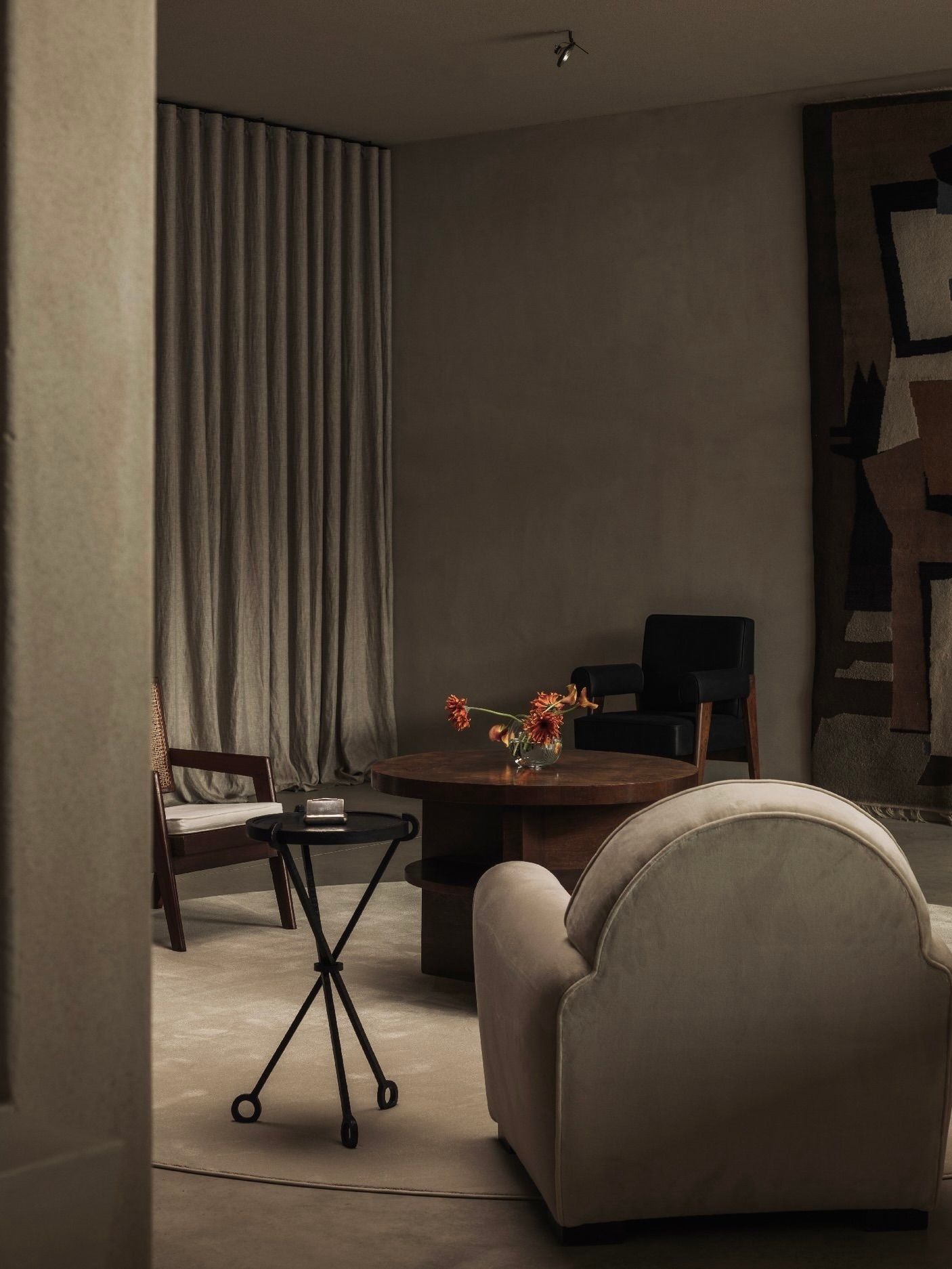 A cozy living room with a neutral palette showcases an armchair, the De Coene Frères Art Deco Guéridon by Bicci de' Medici adorned with flowers, and a black side table. A tapestry decorates the wall, while soft lighting highlights textured drapes, blending elegance and comfort.