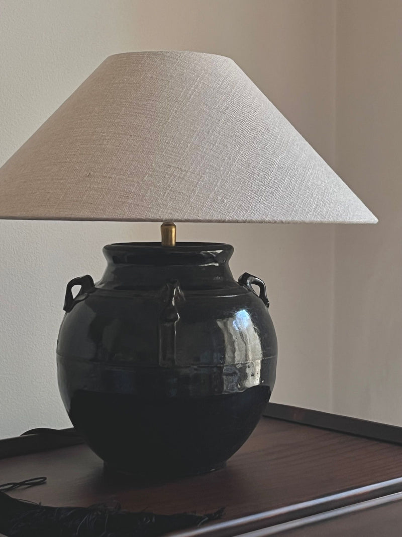 An Antique Glazed Lamp with Ears by Médecine features a glossy dark ceramic vase base with decorative handles, a large off-white conical lampshade, and sits atop a dark wooden surface against a plain light-colored wall.