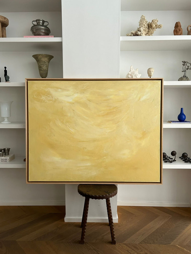 Ethereal Light" by Mattias Storm, an abstract painting with swirling yellow patterns in an oak frame, sits on a wooden stool. It complements the room's white shelves filled with vases, sculptures, and coral. The floor features a herringbone wooden pattern.