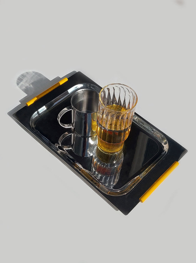 A Les Objoies medium steel tray with two yellow pencils, a stainless steel mug, and a faceted glass half-full of amber liquid, casting a long shadow on a light gray surface.