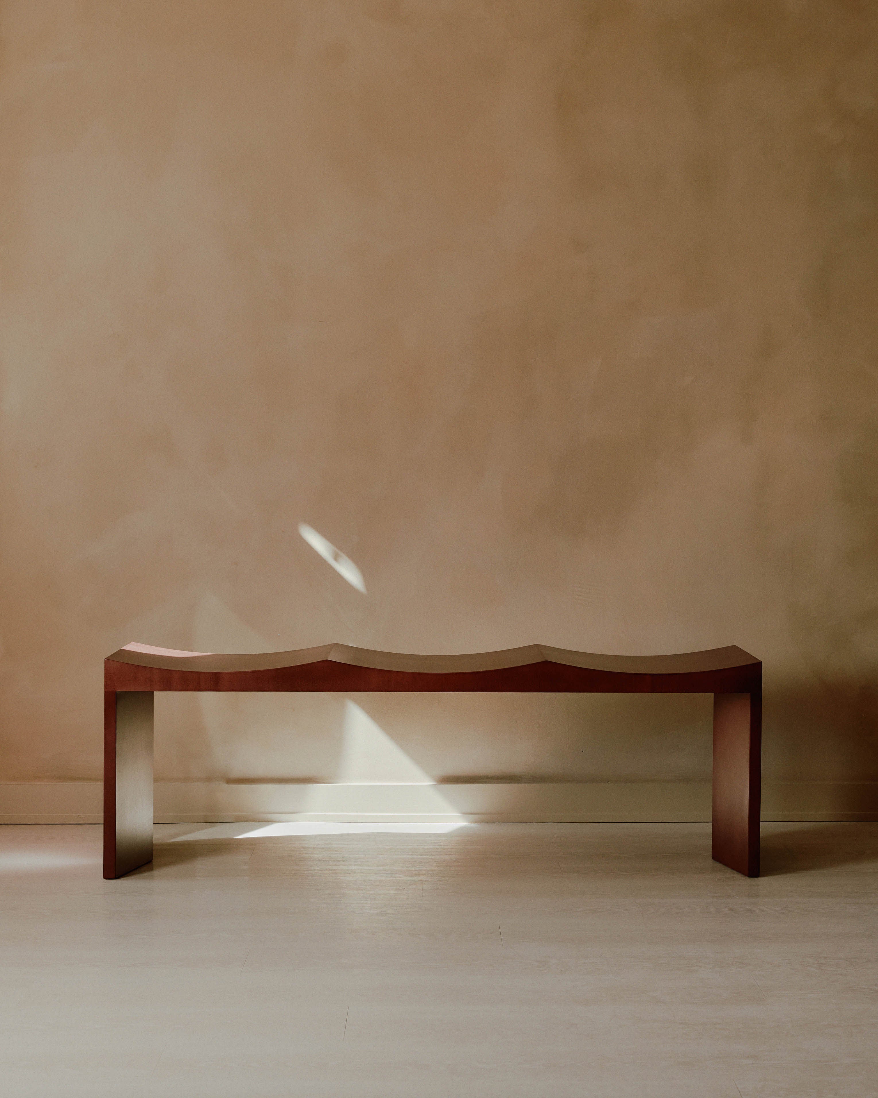 A minimalist Arc Bench by Juntos Projects, featuring a wavy wooden surface, sits in front of a textured beige wall on a light-colored floor. Crafted from premium natural materials, a small beam of light illuminates part of the bench and wall, casting soft shadows.