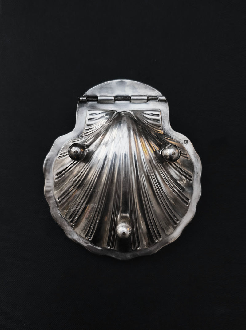 The Silver-plated Shell Box by Les Objoies, a decorative metal container with a clam shape, hinged top, and three knobs, rests against a black background.