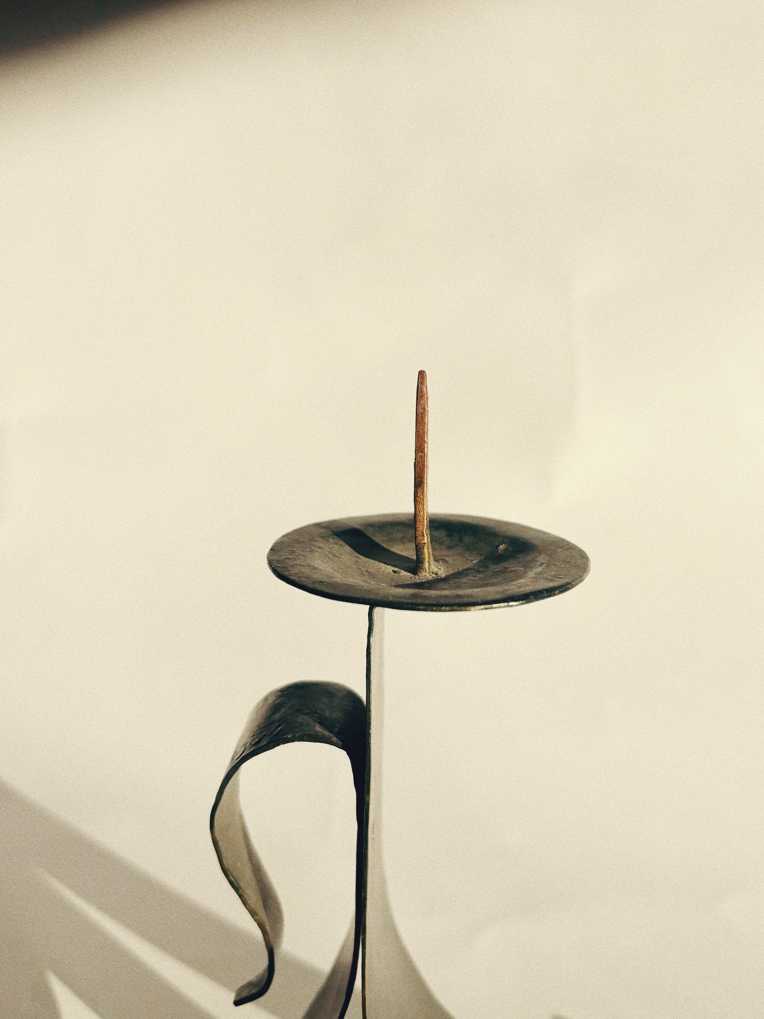 A close-up of Huerto's Handmade Metal Candleholder reveals a slender, curved handle and a central spike without a candle. The softly blurred background accentuates the minimalist design and vintage charm of this artist-made tableware piece.