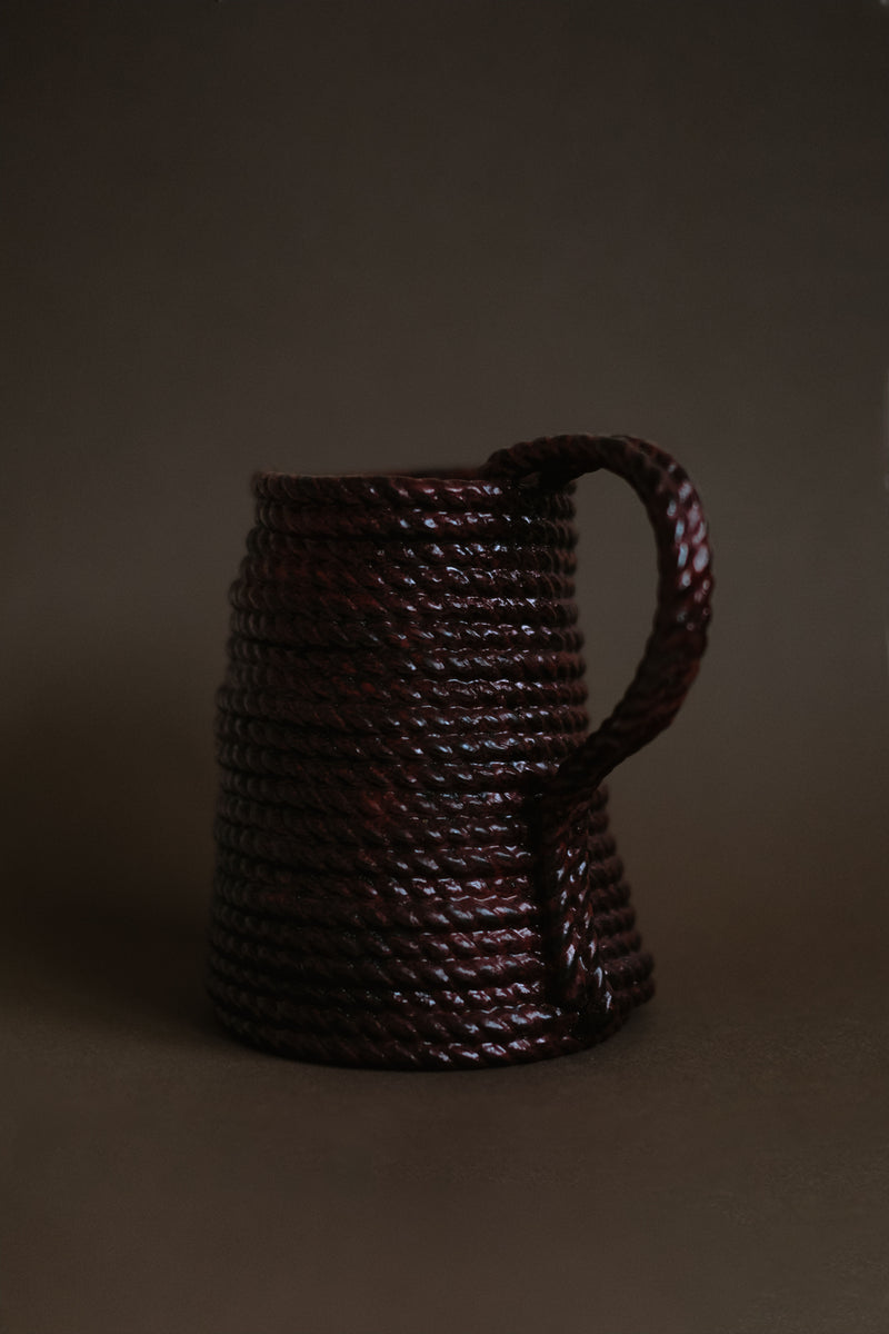 The Handmade Burgundy Vase by septembre studios, featuring a tightly wrapped, glossy rope design in deep brown, rests on a dark surface with a neutral brown backdrop.