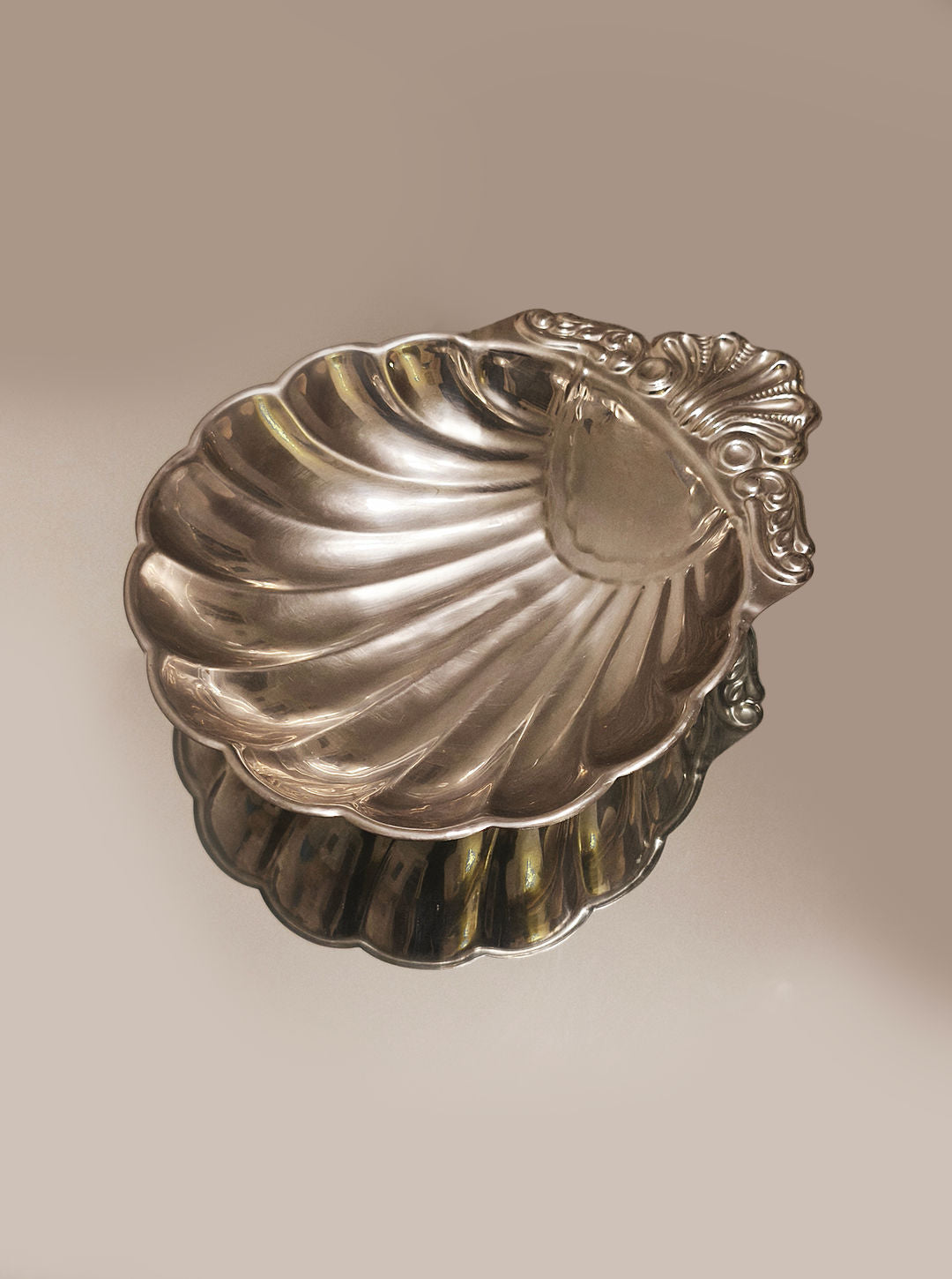 The Les Objoies Large Shell Dish, an ornate shell-shaped dish, features intricate detailing and a polished stainless steel finish that softly reflects light, offering versatility in any setting.