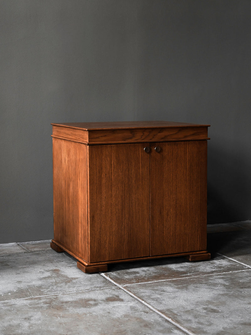 The Lancaster Nightstand by Studio OSKLO, made from stained oak in a rich brown finish with two doors and round metal knobs, rests on a concrete floor against a plain gray wall.