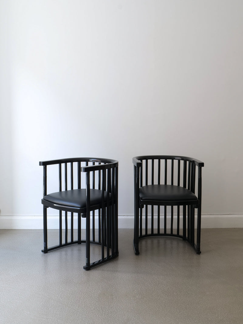 Two sleek and modern Bentwood Armchairs by Collection Apart, featuring Josef Hoffman's signature round backs and vertical slats, are positioned side by side on a smooth, light gray floor. The elegant contrast of their lacquered bentwood against the plain white wall adds a touch of sophistication to the scene.