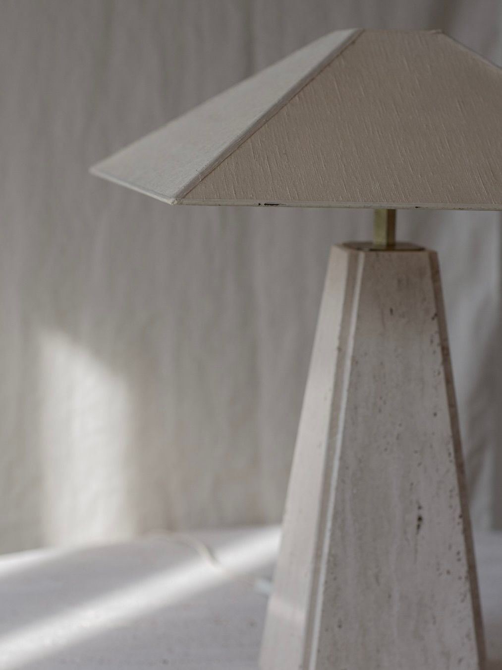 The Bicci de' Medici Camille Breesch Travertine Table Lamp, featuring a cone-shaped shade and textured triangular base, exudes elegance as it stands. Soft natural light casts gentle shadows on the brass surface and background, enhancing its serene appeal.