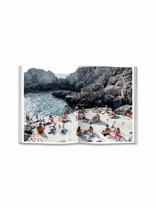 An open copy of the Italian Summer Book by Natalie Obradovich, from Maison Plage, displays a photograph capturing an Italian summer. People are sunbathing on a rocky beach under clear skies, with some swimming in the blue water. The rocky cliffs frame the tranquil seaside atmosphere, showcasing one of the many iconic photos within this beautiful coffee table book.