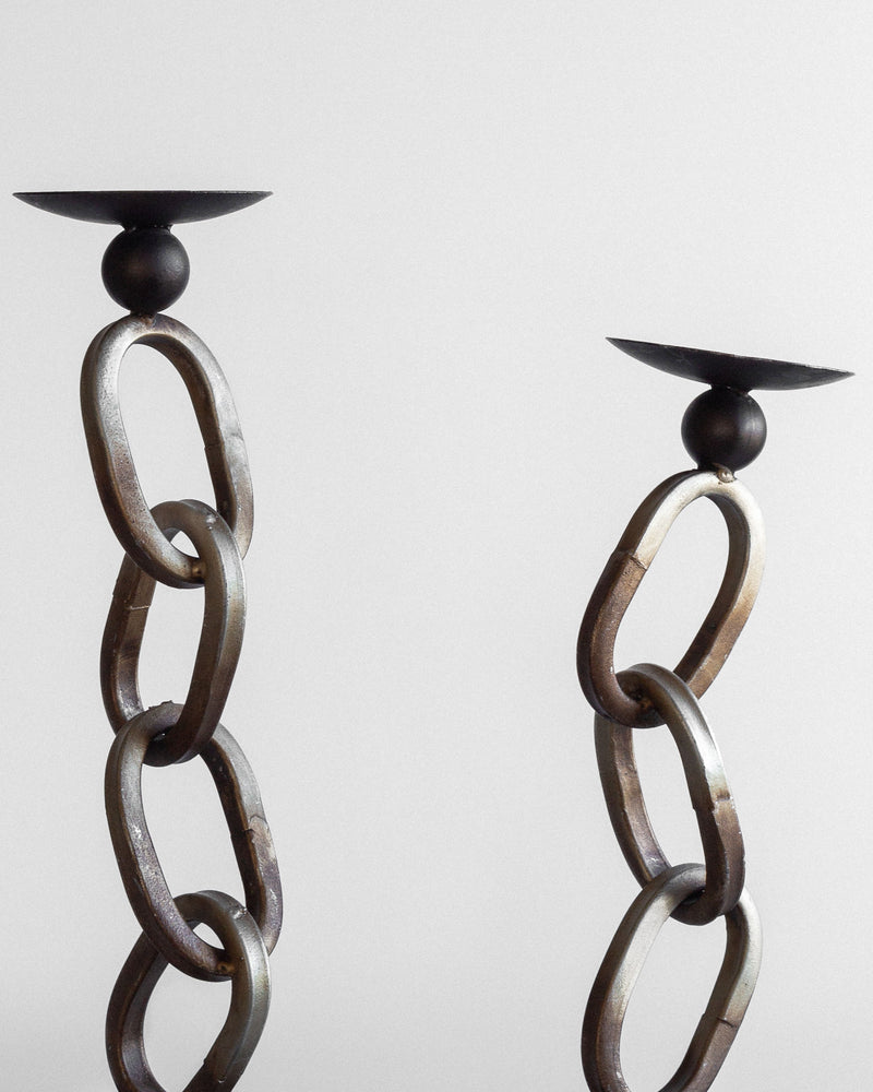 The Postmodern Sculpture Duo from Spigel features two modern candle holders crafted from large interlinked metal rings, embodying an industrial decor style and set against a plain backdrop. The rings, showcasing a rustic appearance in rusty brown, are topped with flat black candle plates. One holder stands taller than the other, creating an asymmetrical visual effect.