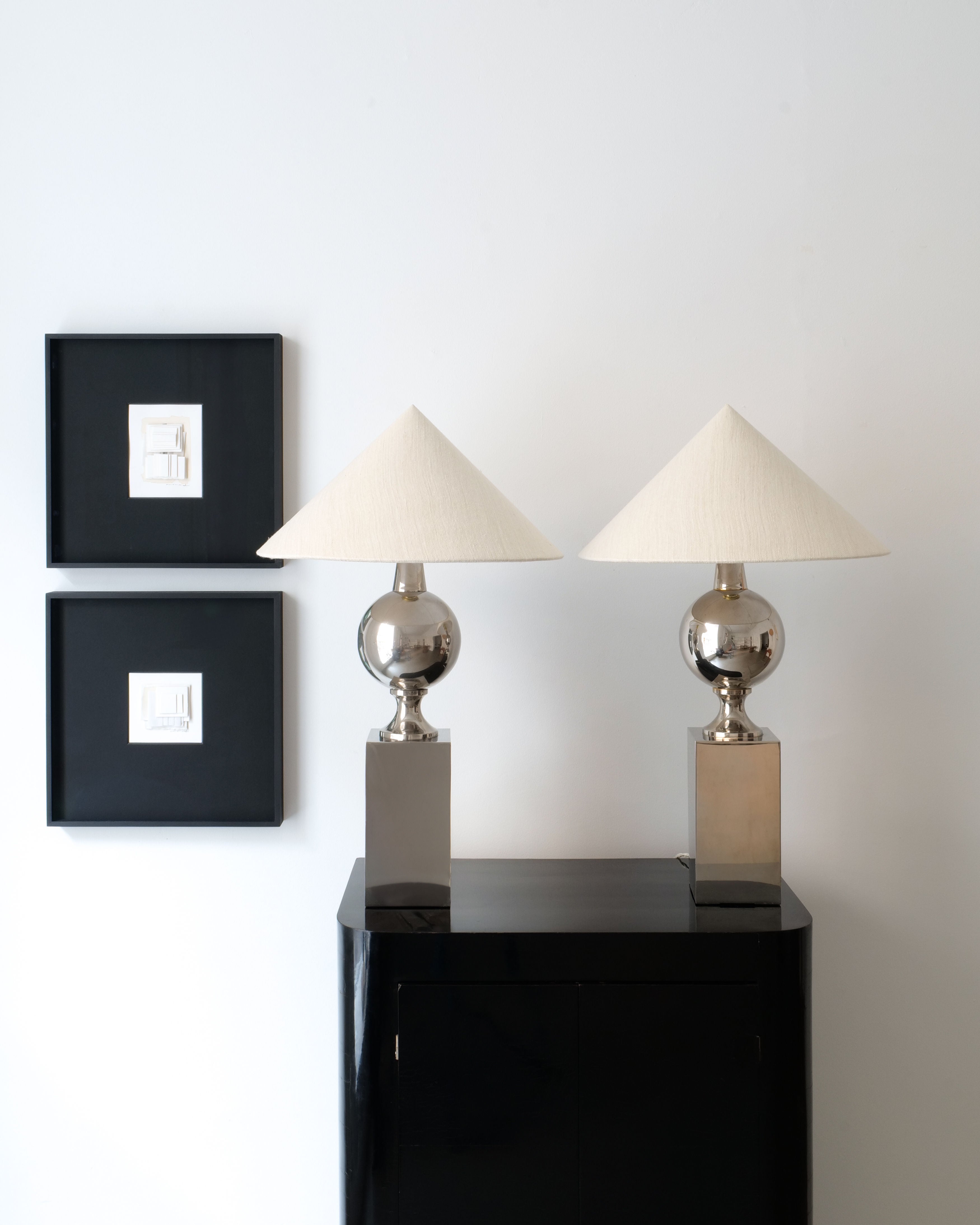 A minimalist interior scene showcases Collection apart's Barbier Chrome Table Lamp with conical shades atop a sleek black cabinet. Two black square frames with white abstract art elegantly adorn the white wall in the background, enhancing the sophisticated ambiance.