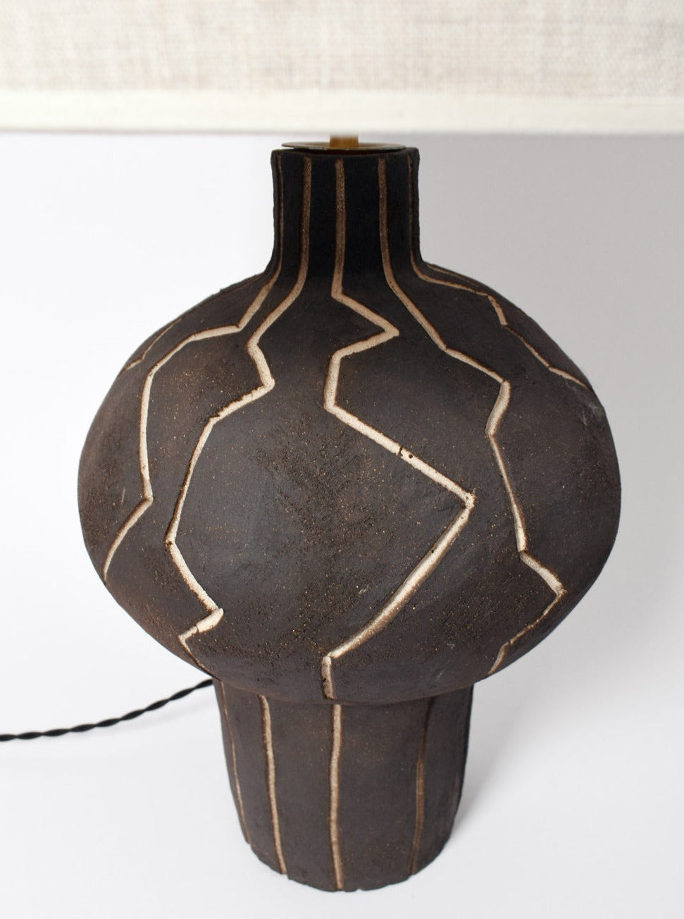 A modern ceramic table lamp featuring a rounded, vase-like shape with a dark matte finish. Made in Portugal, this black "Lanzarote" Lamp by Barracuda Interiors showcases an abstract linear pattern etched into its surface, exposing a lighter color underneath. The lamp is switched off and the power cord is partially visible.