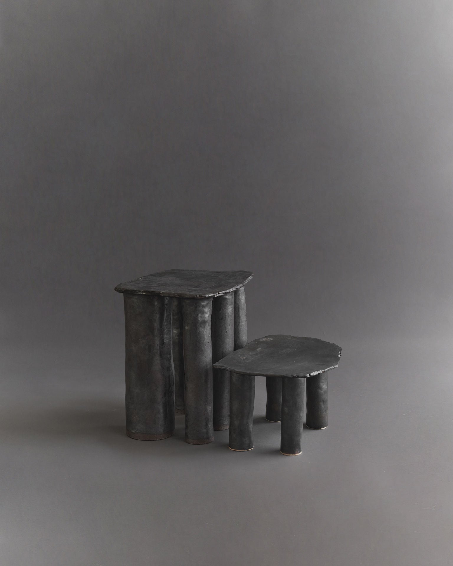 The Cleo Ceramic Side Tables by Ombia Studio are two uniquely shaped dark tables with irregular forms. The larger features six cylindrical legs, and the smaller has five, both showcasing a textured black glaze surface reminiscent of handmade craftsmanship against a plain gray background.