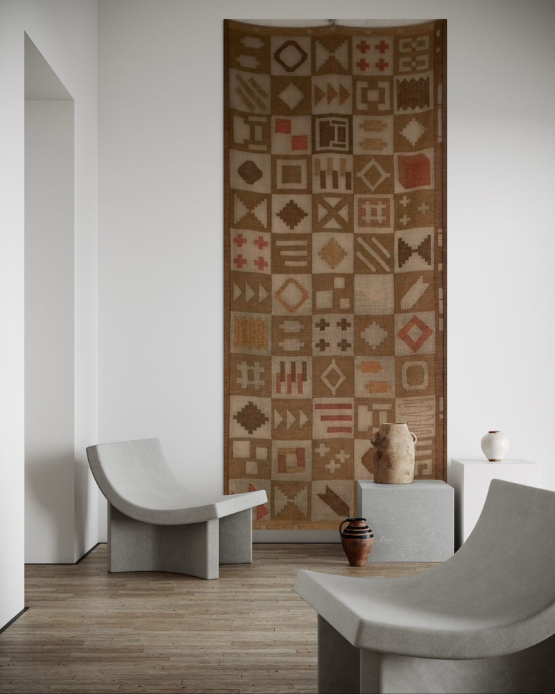 A minimalist room features curved concrete chairs, wood flooring, neutral pottery on gray blocks, and Veter Vintage’s Large Abstract Wool Wall Tapestry (Sweden, 1988) with geometric motifs displayed vertically on a white wall.