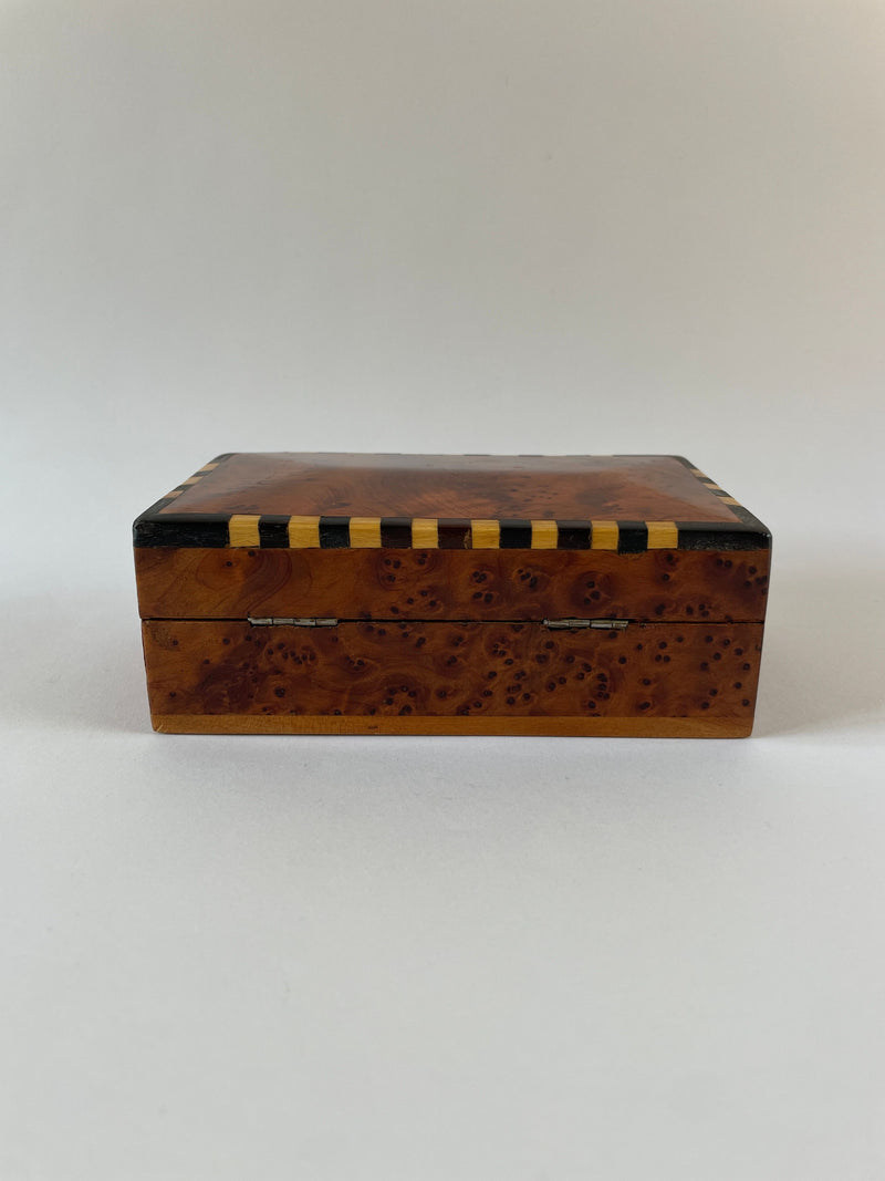 Introducing the Art Deco Burr Box by Maud Vaughan, a meticulously handcrafted wooden box featuring a glossy finish and distinct art deco style. The lid is adorned with alternating black and yellow stripes along the edges, beautifully contrasting the rich burr veneer that enhances its dynamic grain. Its background presents a simple, neutral tone.