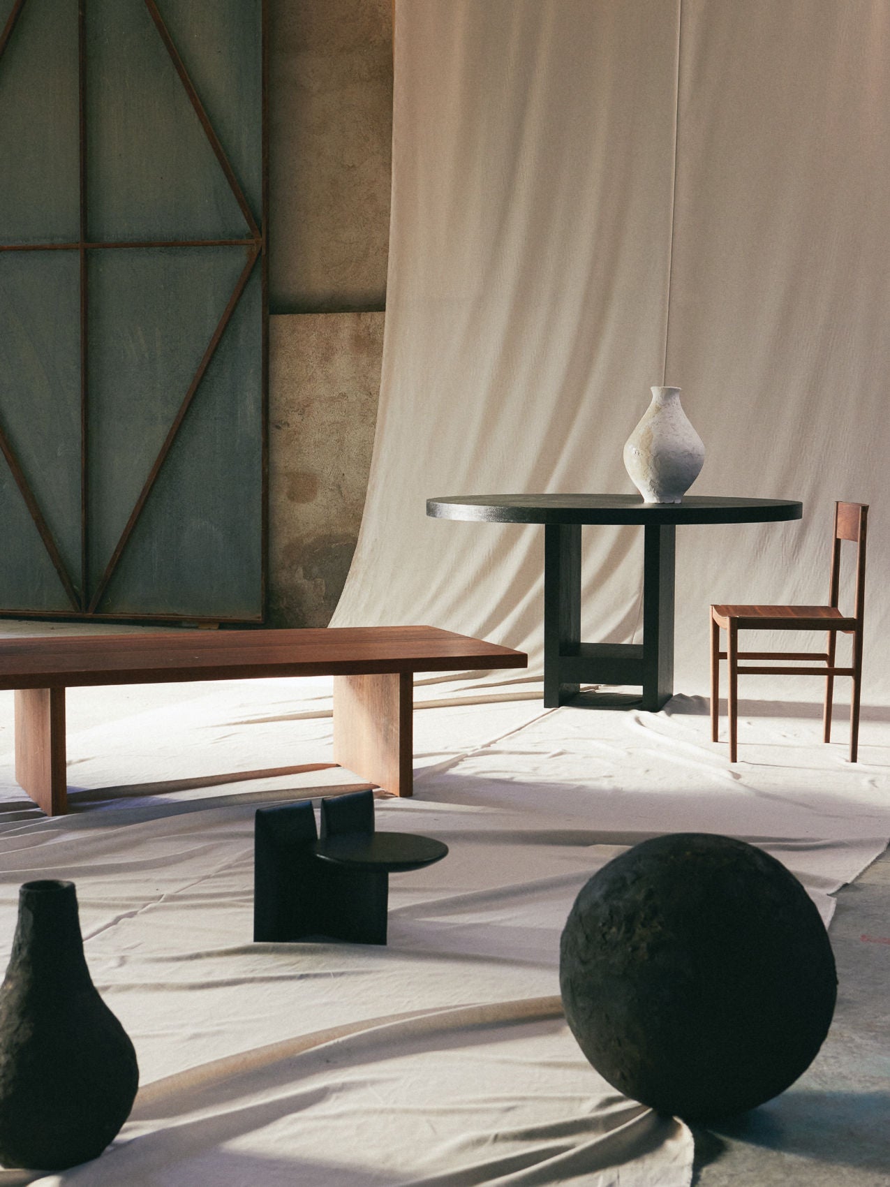 The minimalist room showcases a Sister Table by ASERIES OF OBSTACLES, SL, paired with a solid oak chair and large vase. Abstract sculptures adorn the floor, while geometric-patterned backdrops enhance the artistic atmosphere, seamlessly blending natural materials for warmth and charm.