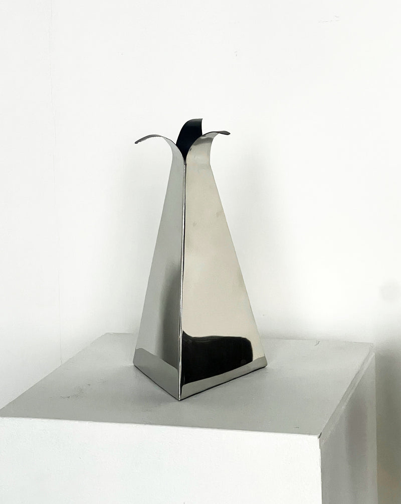 The Triangular Vase by Porter Studio is a sleek, metallic styling piece shaped like a tall triangle. It features three pointed, outward-curving tips and stands elegantly on a white pedestal against a plain white background.