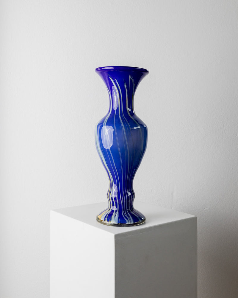 A tall, glossy Murano Glass Vase Blue by Bottega Jacobs from the 1970s, featuring white marbling, stands on a white pedestal against a gray background, showcasing exquisite Italian glass art.
