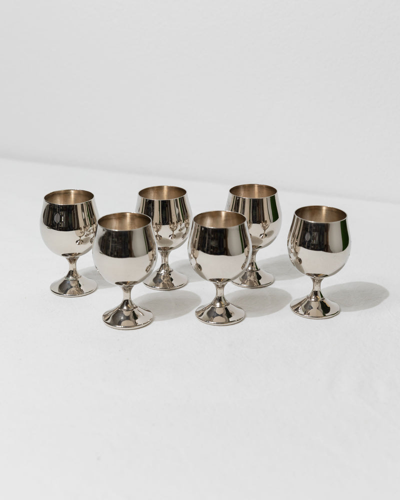 Six silver-plated eggcups from Bottega Jacobs are arranged on a white surface, evenly spaced with reflections on their polished surfaces, creating a minimalist and elegant composition that evokes a mid-century design reminiscent of 1980s Italy.