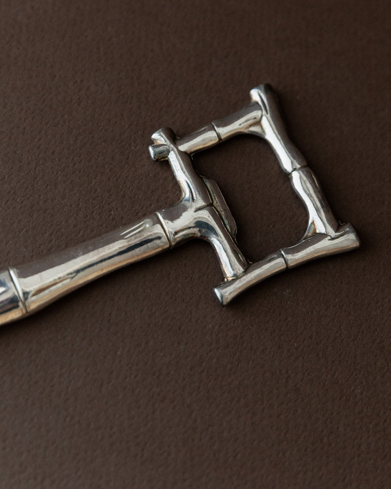 A silver-plated 1970s Bottle Opener by Bottega Jacobs, featuring a complex double-bow design reminiscent of a skeleton key and Hollywood Regency style, rests on a dark brown surface with intricate details and a shiny finish.