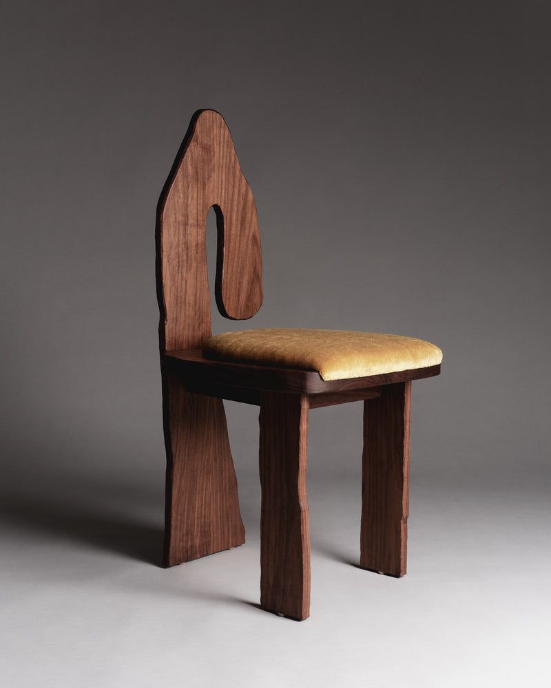 The Muisca Dining Chair by Ombia Studio is a solid oiled walnut chair with an abstract backrest and uneven, sculpted legs. The seat is upholstered in yellow mohair fabric, set against a plain dark gray background for a mix of artistic flair and comfort.