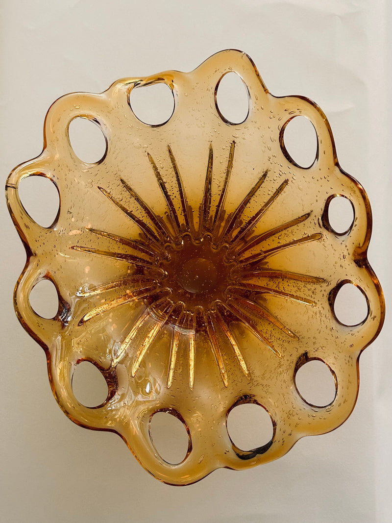 The Murano Glass Amber Bowl by Flock Studio showcases Italian glass artistry, with scalloped edges and circular cutouts along the rim. Its textured center, reminiscent of a sunburst, enhances its retro charm as it rests on a light surface.