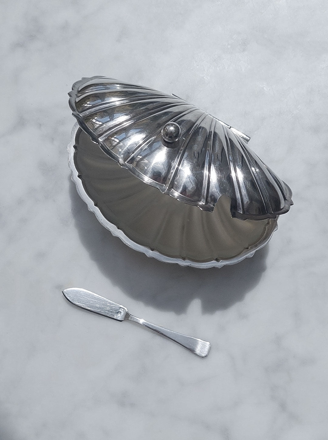 A closed, Les Objoies Shell Silver-Plated Butter Dish with a seashell shape and a small knob on top sits on a marble surface. A matching silver butter knife rests beside the dish. The reflective surfaces of both items give a polished, elegant appearance, exuding vintage charm.