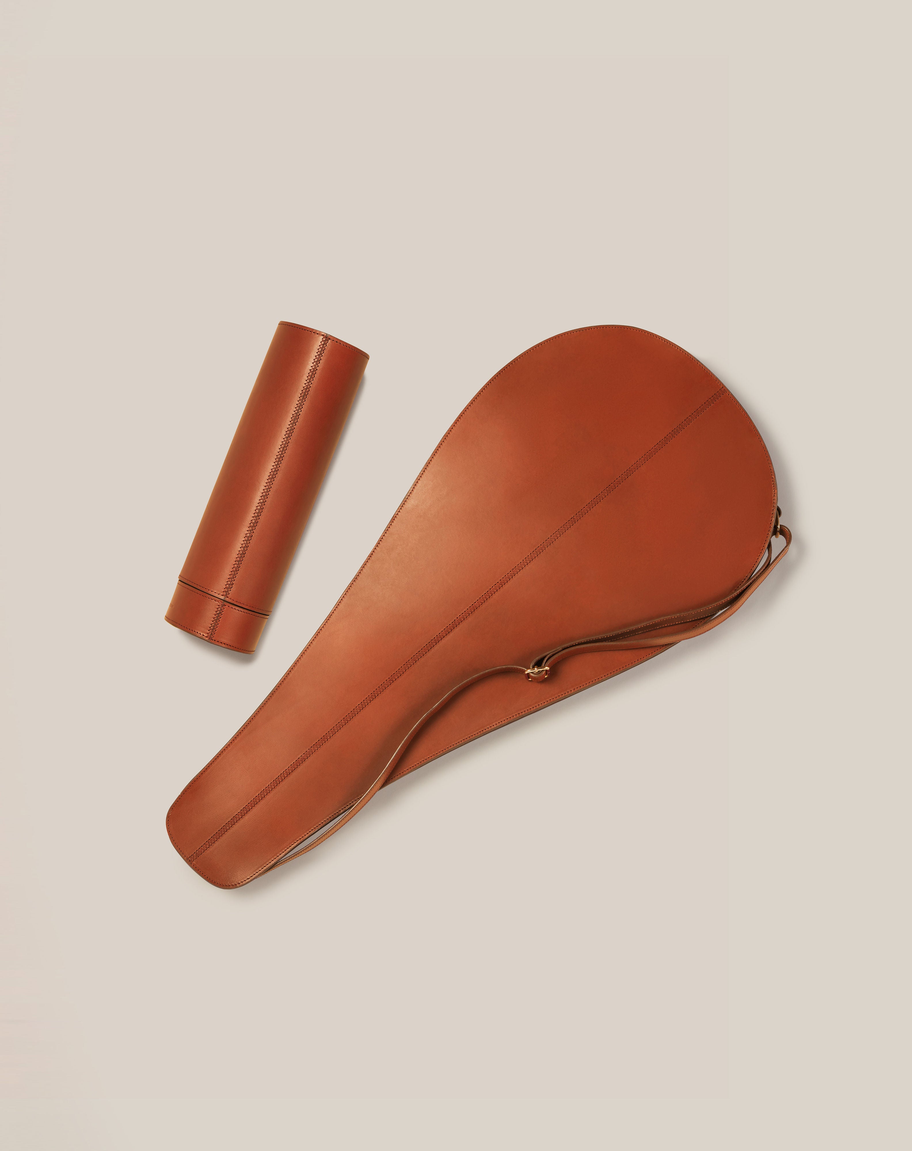 A Paradise Row brown leather tennis racket cover, made from sustainable vegetable-tanned leather, rests beside a matching smooth cylindrical container on a light beige background. The bespoke British design includes a zipper and handle on the cover, with the container featuring a cap.