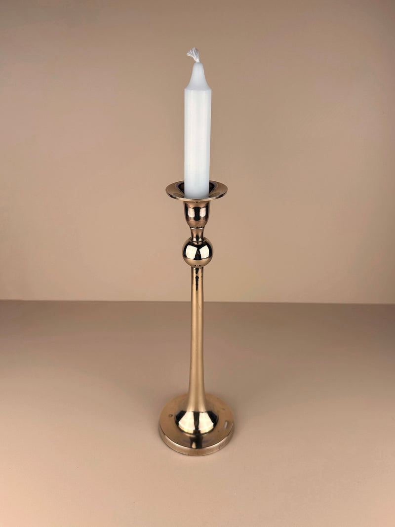 A tall, elegant large candlestick with a white, unlit candle stands on a beige surface. The Dodo Vintage Postmodern Brass Candleholder features a reflective surface and a classic, slender design. The background is a soft, neutral tan color, complementing the polished brass of the Dodo Vintage Postmodern Brass Candleholder.