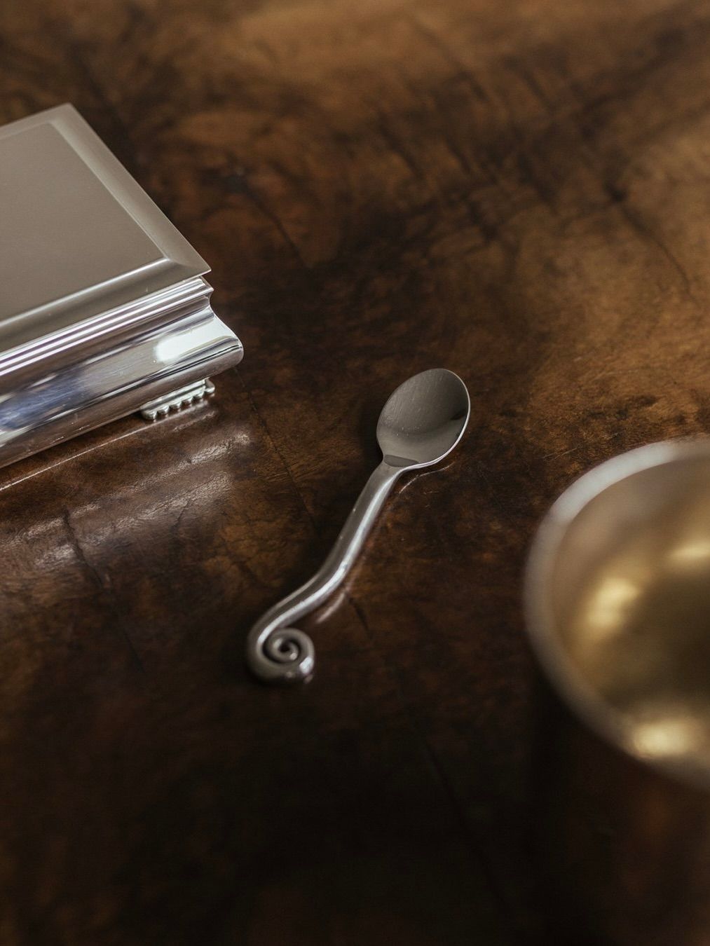 A small metal spoon with an ornate, curled handle rests on a polished walnut burl veneer table. Nearby, a metallic box with a decorative edge sits next to the De Coene Frères Art Deco Guéridon by Bicci de' Medici, where the sheen of a cylindrical metal cup is just visible.