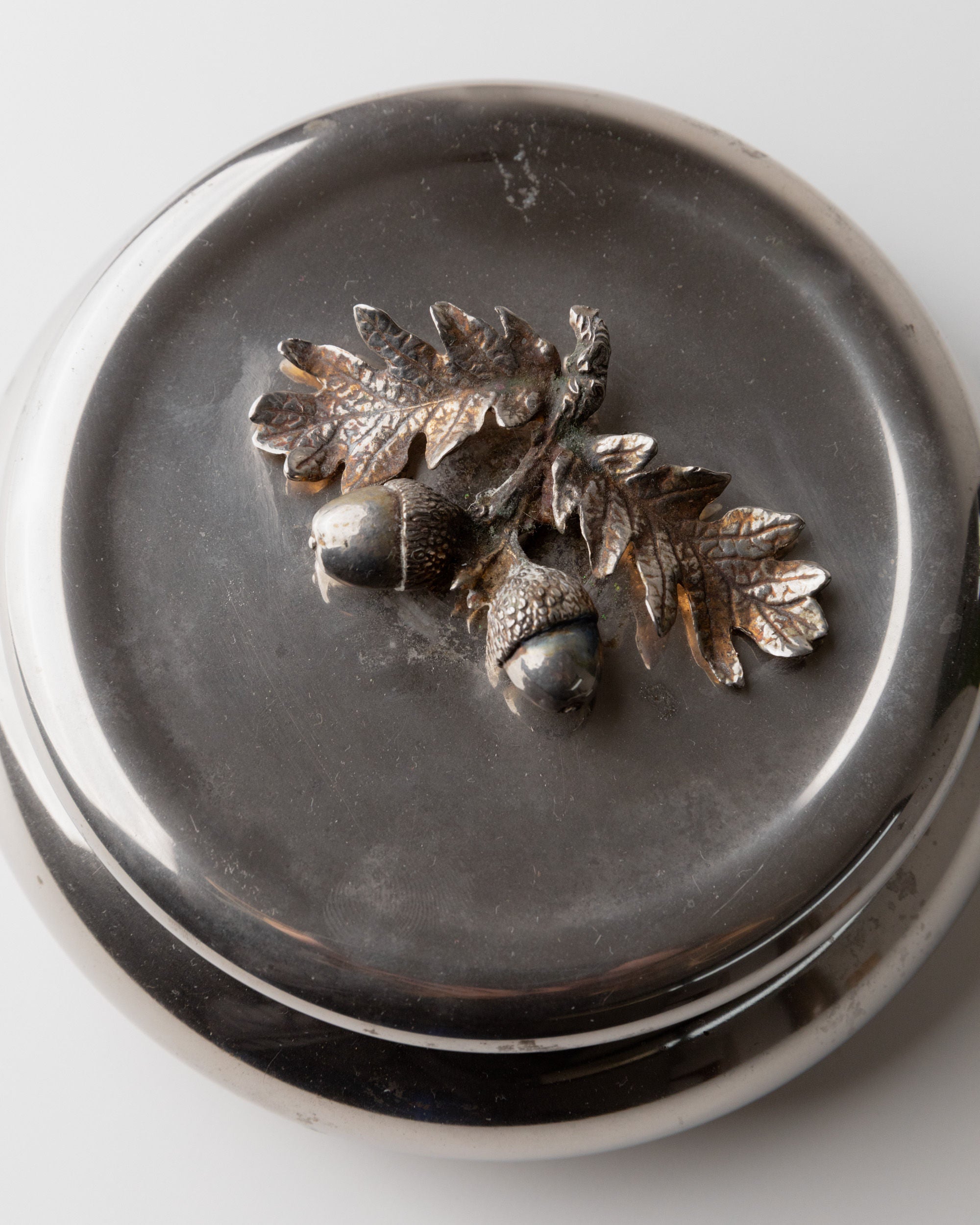 A round, silver-plated box by Bottega Jacobs, from 1950s Italy, features an antique finish with a decorative relief of two acorns and oak leaves against a smooth background, resembling an elegant vintage tin.