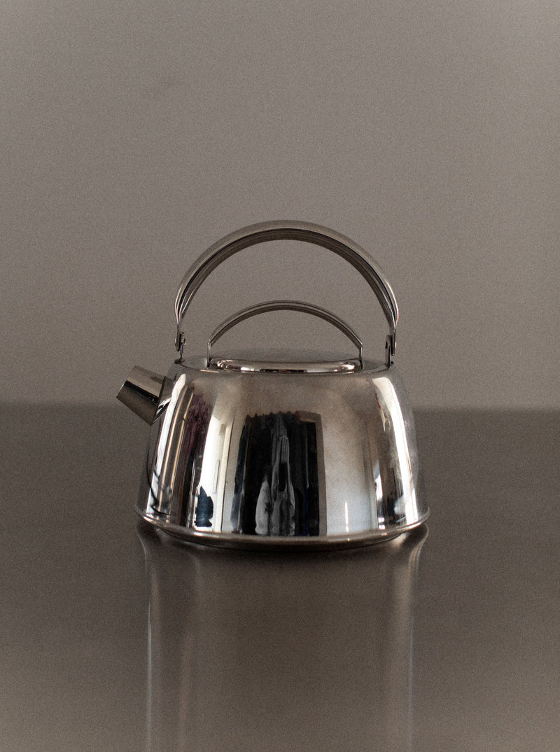 An Out For Lunch Chrome Teapot with a handle and spout sits on a reflective surface. The teapot has a polished, shiny finish that reflects parts of its surroundings, adding to its aesthetic appeal. The background is neutral and uncluttered, enhancing the refined dining experience.
