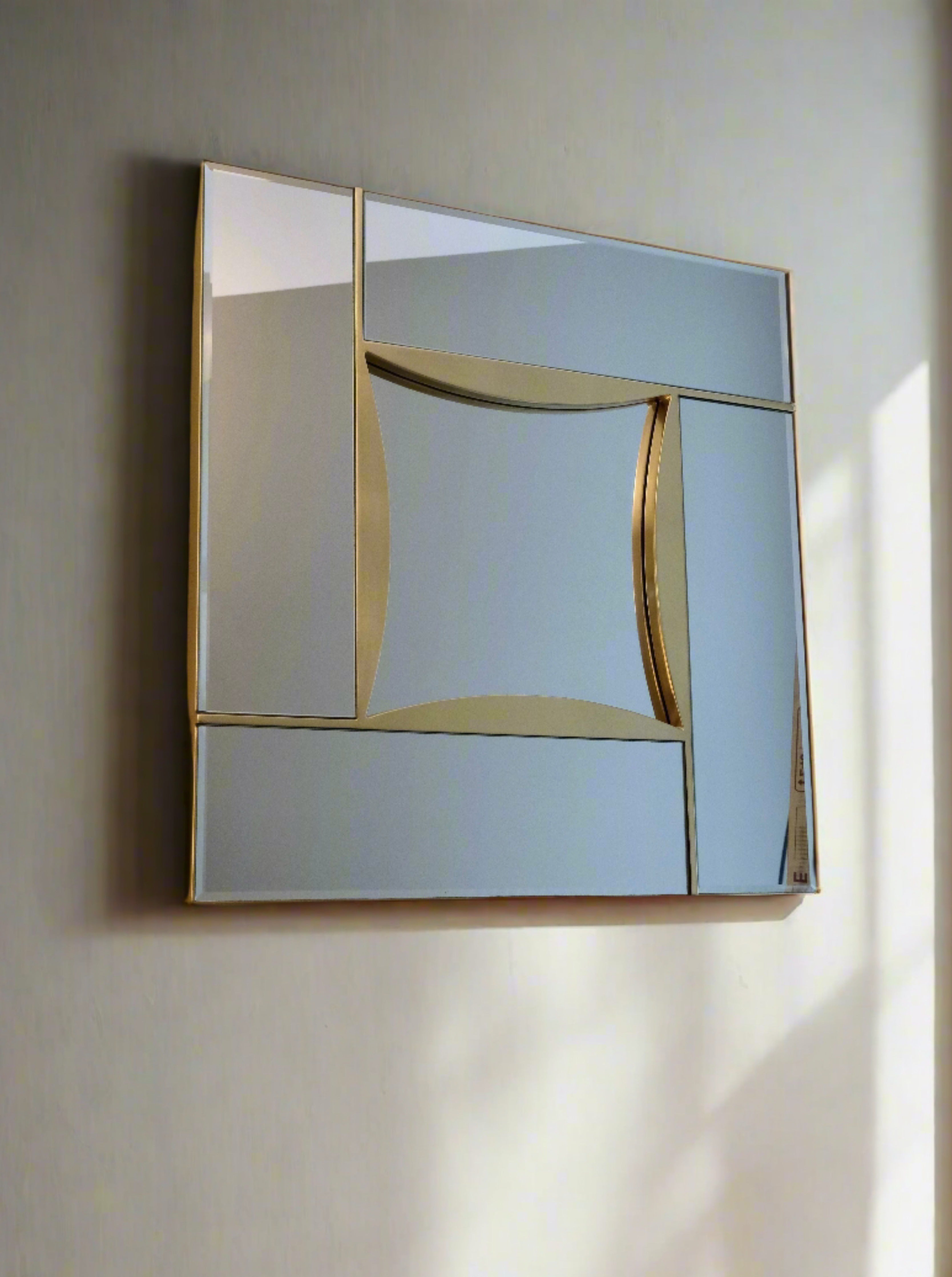 A modern rectangular wall mirror with an abstract design featuring a combination of mirrored and gold sections. Mounted on a light-colored wall, the Mirror Belgo Chrome in Beveled Glass and Brass by LA NOUVELLE GALERIE includes cloisonné glass as a decorative element. Sunlight casts a soft shadow, adding depth and contrast to the scene.