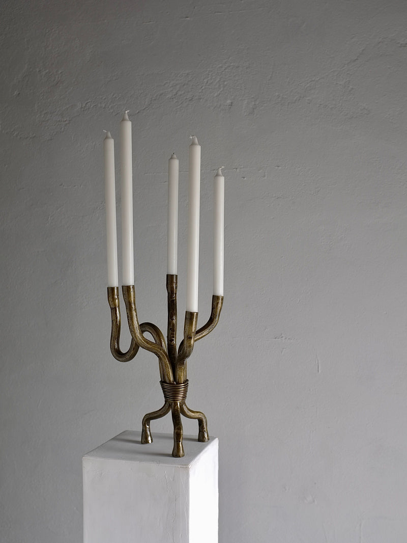 A Veter Vintage "Brutalist Brass Candelabra" from the 1970s Netherlands, featuring an artistic twisted design, stands in vintage condition on a white pedestal with six tall, unlit white candles against a plain, textured gray wall.