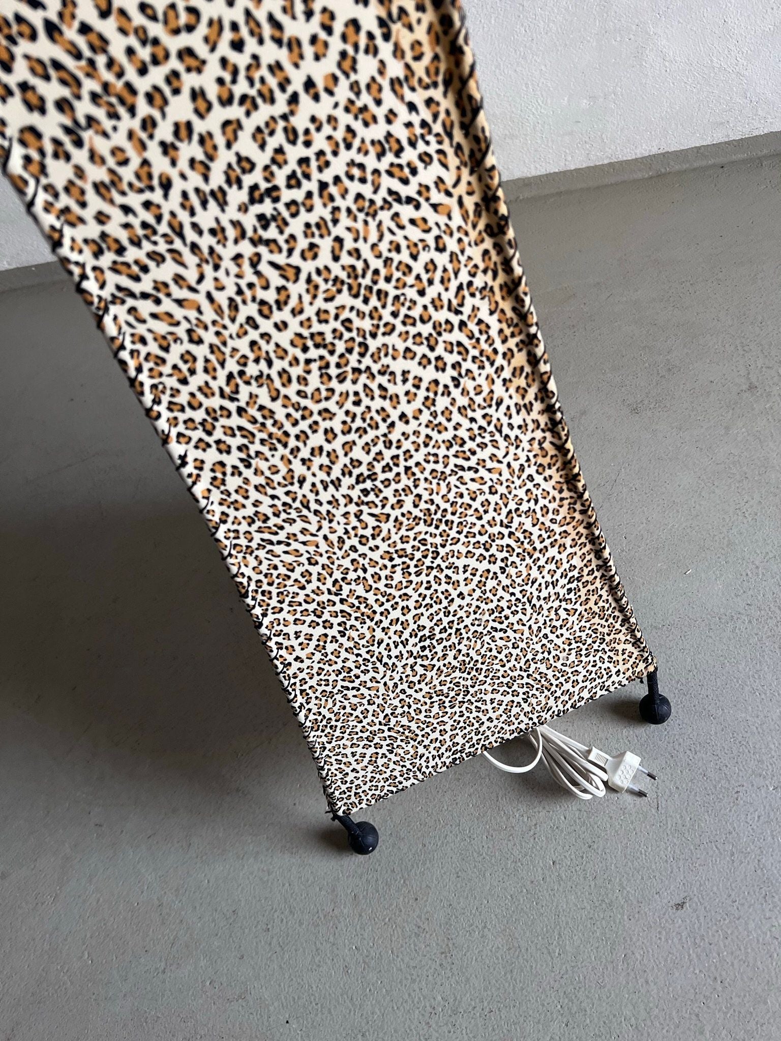 The Veter Vintage Leopard Print Floor Lamp from the 1980s features a slim, vertical design on four small wheels, with power cords coiled at its base. The leopard print cover adds a retro touch to any gray-floored space.