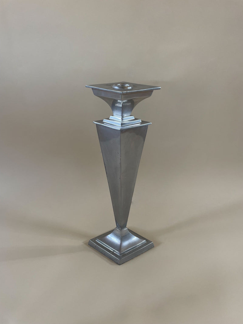A tall, silver candlestick from the Maud Vaughan Pair of French Art Deco Candleholders collection stands on a beige surface. This metallic piece, reflecting the characteristic French Art Deco style, features an angular geometric design with a square base and holder at the top and is highlighted by a tapered inverted pyramid-shaped body in the center.