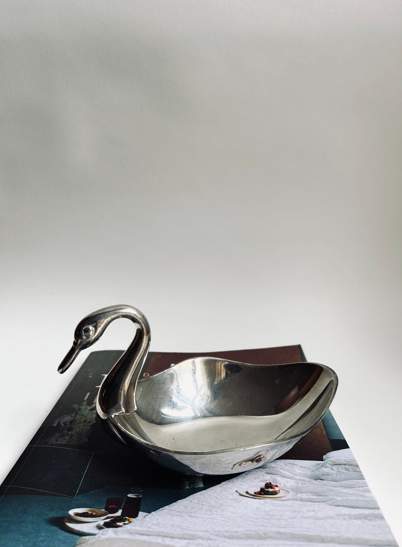 A reflective Silver Plated Swan Dish by Flock Studio rests on an open magazine known for its party decor inspirations, showcasing a photo of a white bedspread adorned with small garnishes and accompanied by two glasses of wine to create a luxurious and serene ambiance.