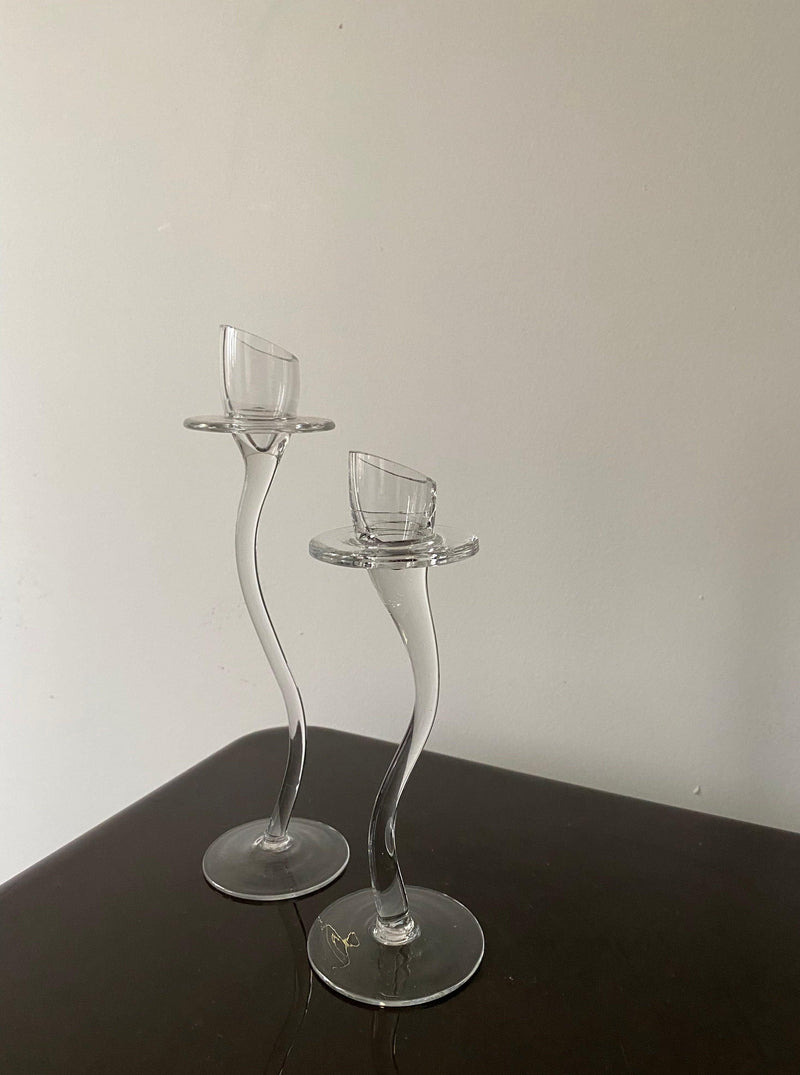 Two tall, slender Les Objets Amsterdam vintage-inspired glass candle holders with intricate stems, placed on a dark wooden surface against a plain, light-colored wall.